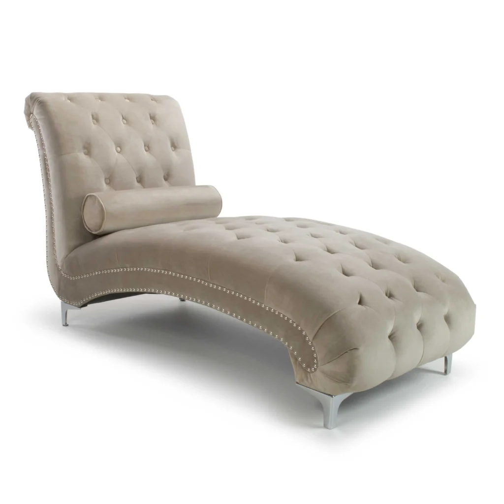 Elegant beige velvet chaise lounge with tufted upholstery and nailhead trim. Features a curved design and cylindrical pillow, on shiny metal legs.